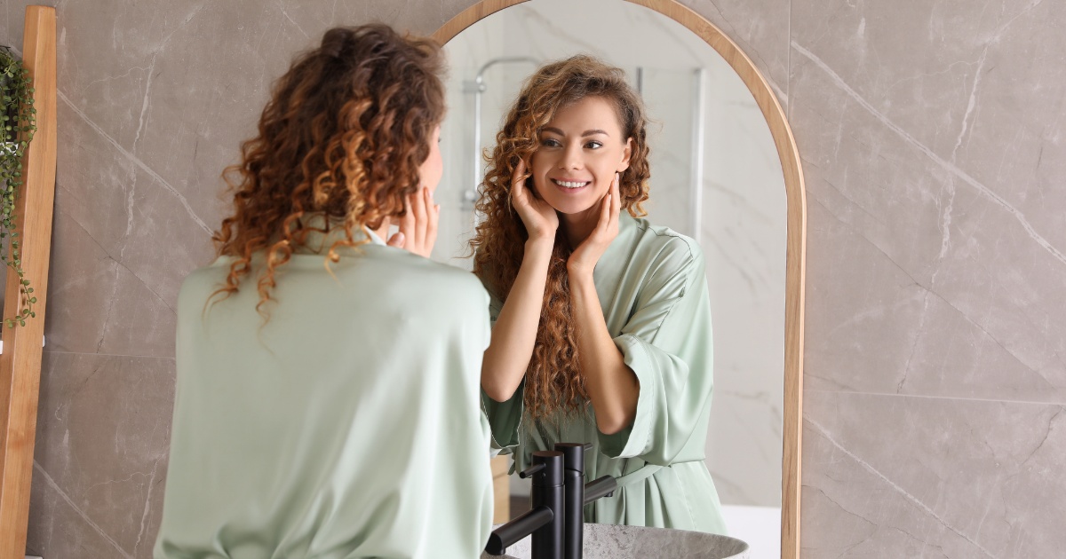 Woman in a light green robe smiling at her reflection in a bathroom mirror, gently touching her face, conveying self-care, confidence, and wellness.
