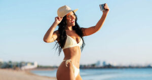 A person wearing a striped bikini and straw hat takes a selfie on a sunny beach, smiling with the ocean and a clear blue sky in the background.